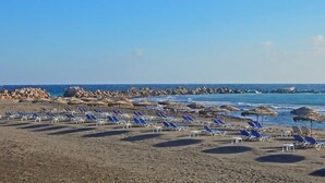 Een strand met rijen ligbedden en parasols, een rotsachtige kustlijn en een helderblauwe lucht.