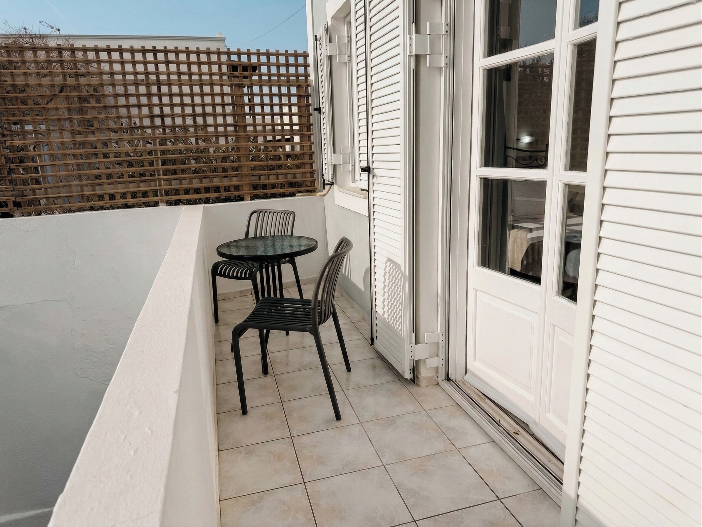 Een klein terras met een ronde tafel en twee stoelen, een houten hek op de achtergrond, en een wit gebouw met een raam.