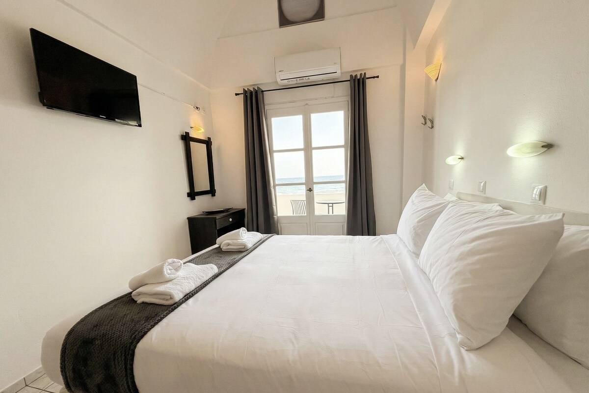 Ein Hotelzimmer mit Bett, Flachbildfernseher, Spiegel, Nachttisch und Meerblick durch ein Fenster.