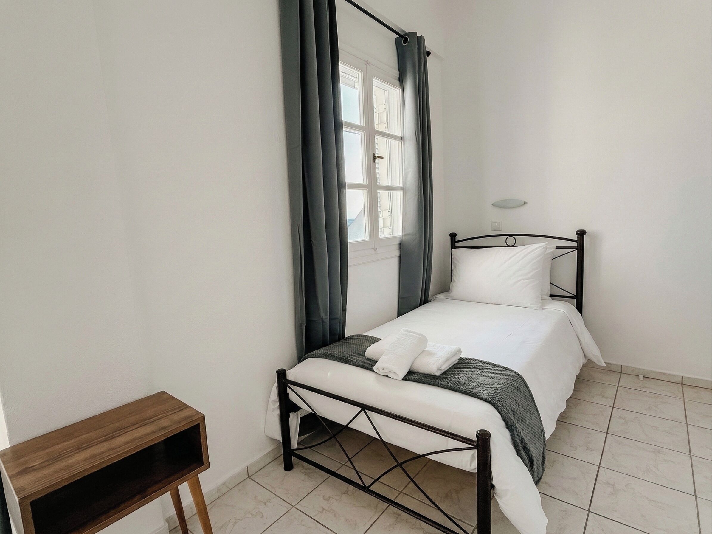 A small, neatly arranged bedroom with a single bed, a bedside table, and a window with curtains.