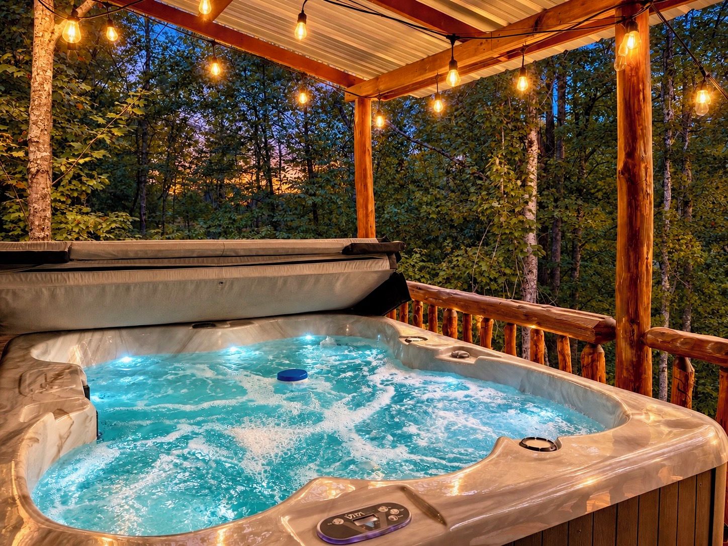 A hot tub with a control panel, surrounded by a wooden deck and string lights, set against a backdrop of trees.