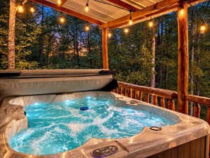 Un bain à remous doté d’un panneau de commande, entouré d’une terrasse en bois et illuminé de guirlandes lumineuses, le tout se fondant dans un décor de verdure.