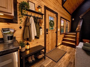 Un intérieur en bois avec un porte-manteau, une étagère remplie de livres, un banc où sont posées des chaussures et une porte ornée d’une guirlande.