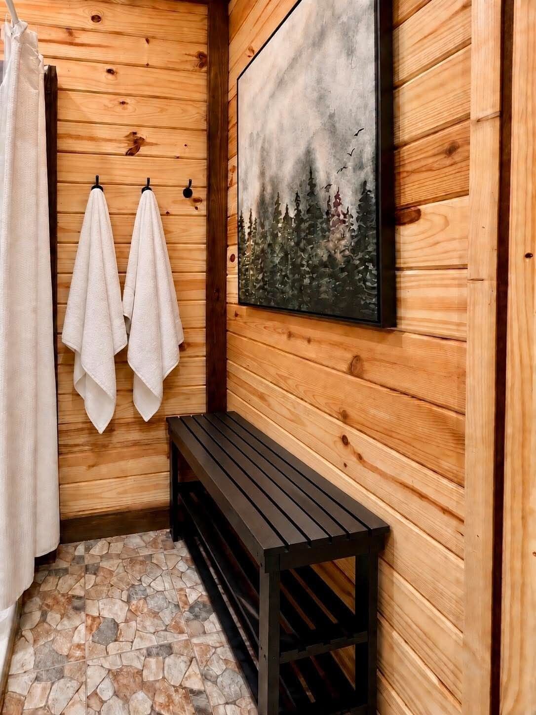 A wooden cabin bathroom with a bench, two towels hanging, a shower curtain, and a painting of trees.