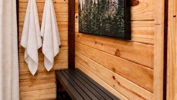 A wooden cabin bathroom with a bench, two towels hanging, a shower curtain, and a painting of trees.