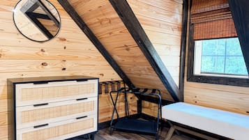 A room with a sloped ceiling, wooden walls, a bench, a dresser with drawers, a window with blinds, and a circular mirror.