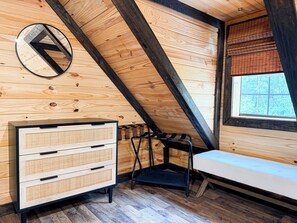 Une pièce avec un plafond en pente, des murs en bois, un banc, une commode avec des tiroirs, une fenêtre avec des stores et un miroir rond.