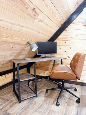 Un bureau en bois, sur lequel se trouvent un écran d’ordinateur, une lampe et une chaise en cuir, dans une pièce aux murs en bois inclinés.