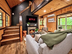 A cozy cabin interior with a fireplace, a Netflix TV, and a staircase leading to a loft. - Cozy Winter Cabin: Steamy Hot Tub, Fire Pit, Game Room, Family + Pet Friendly (Campton)