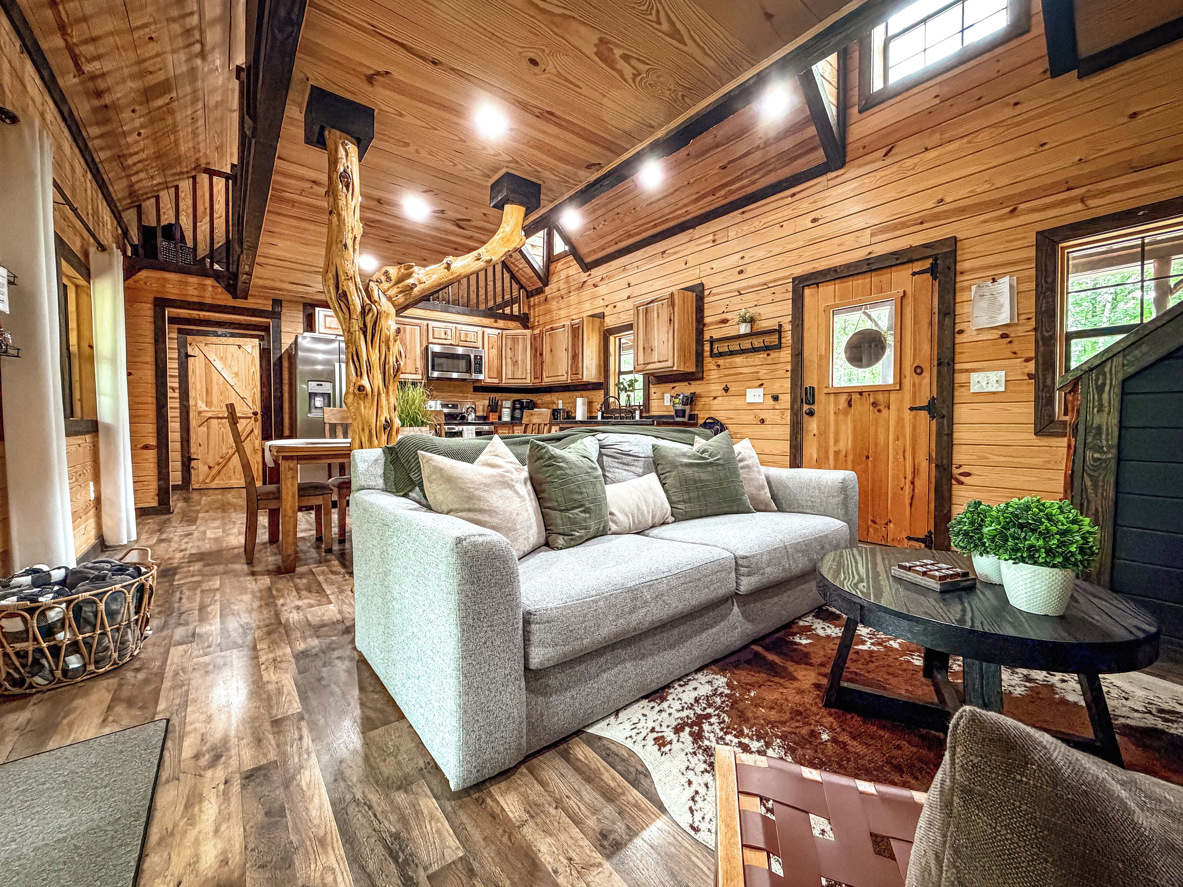 A cozy cabin interior with a wooden ceiling, a sofa with cushions, a dining table, and a kitchen area.