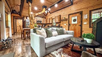 A cozy cabin interior with a wooden ceiling, a sofa with cushions, a dining table, and a kitchen area.