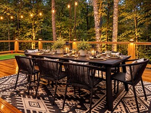 Un espace de restauration en plein air avec une table dressée pour six personnes, entourée de chaises en osier, sur une terrasse en bois agrémentée d’un tapis à motifs.