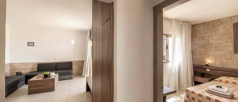 A modern hotel room with a sofa, a wooden coffee table, a bed with a checkered bedspread, and a bathroom visible through an open door.