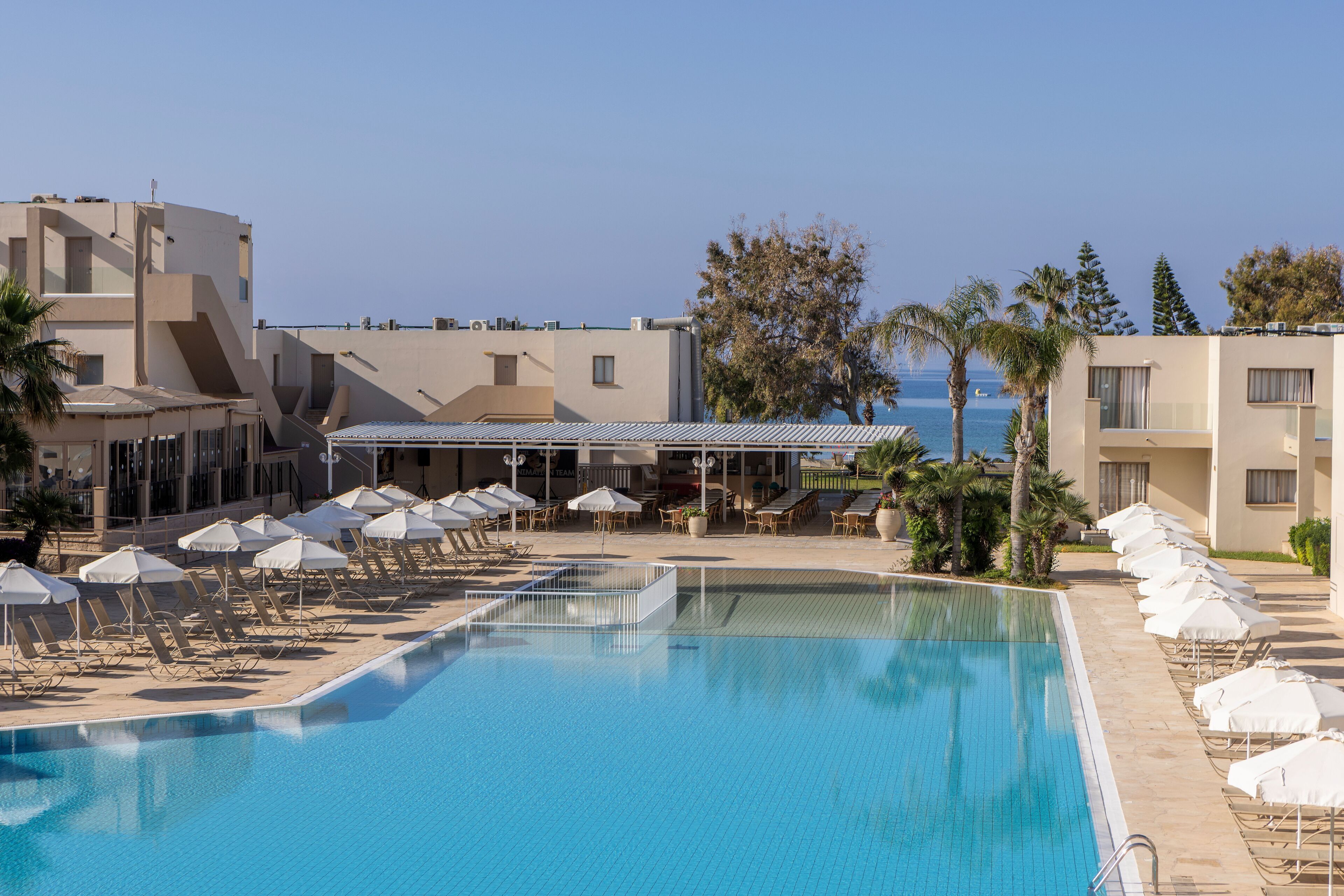 A swimming pool with lounge chairs and umbrellas, a clear sky, and a building with a flat roof.