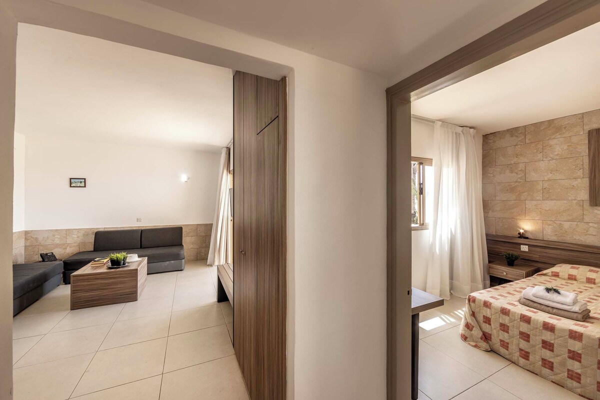 Egy modern szállodai szoba, amelyben van egy kanapé, egy fa kávézóasztal, egy kockás ágytakaróval borított ágy, és egy fürdőszoba, amely egy nyitott ajtón keresztül látható.