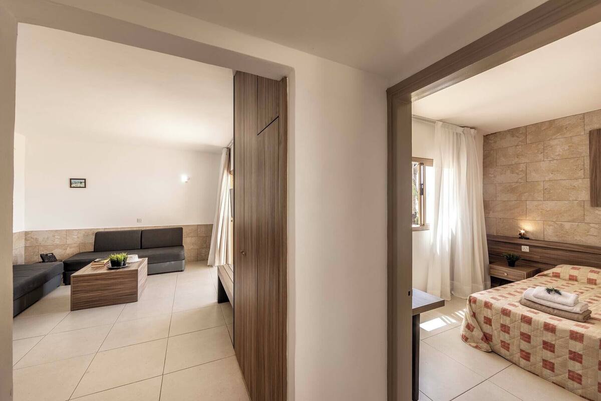 Een moderne hotelkamer met een bank, een houten bijzettafeltje, een bed met een geruit dekbed en een zichtbare badkamer door een open deur.