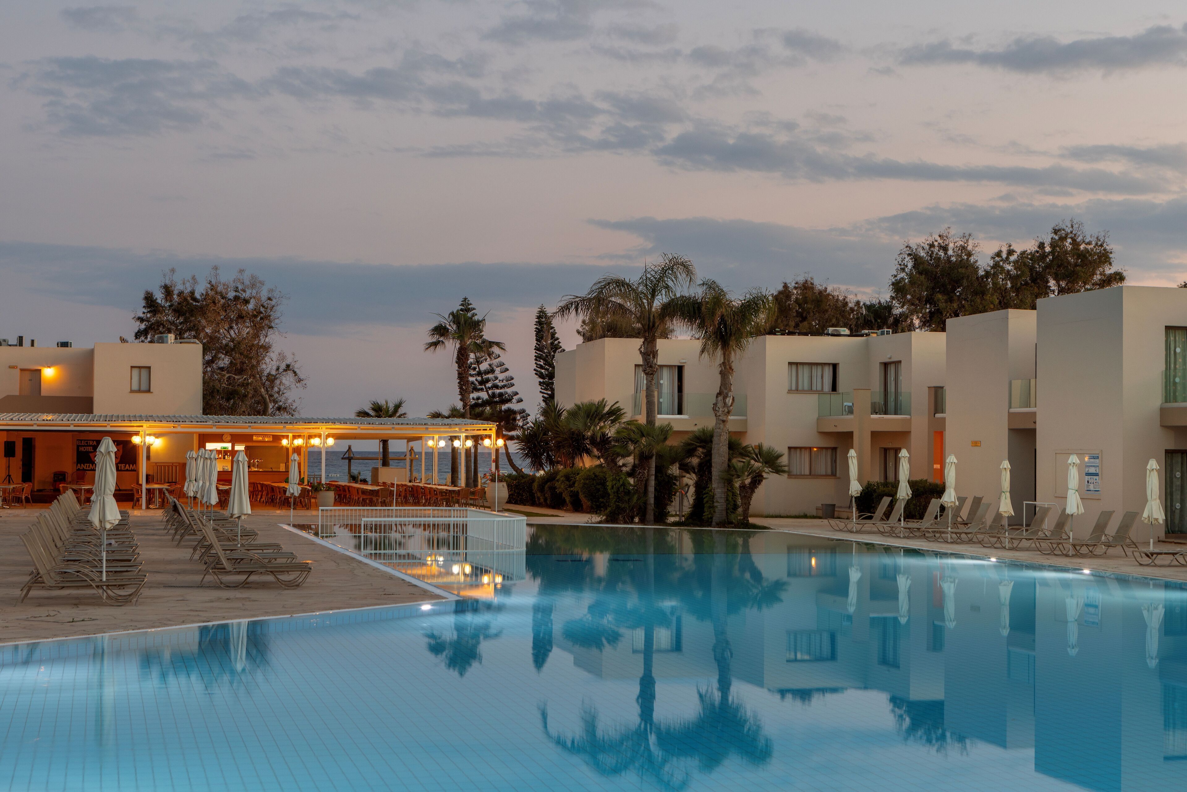 Una piscina con sillones de descanso y sombrillas, un edificio de varios pisos y palmeras.