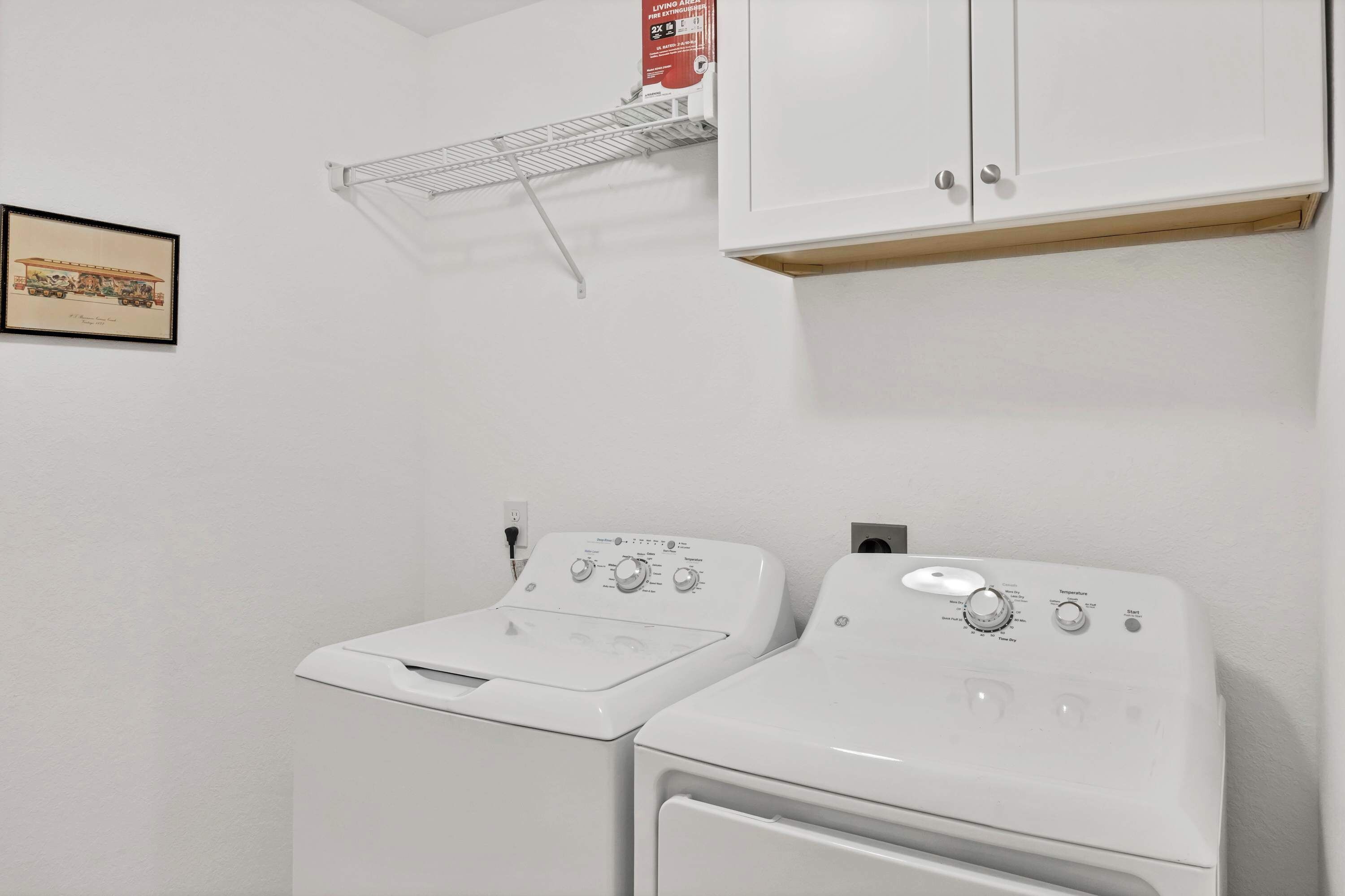 A laundry room with two white washing machines, a metal drying rack, and a framed picture on the wall.