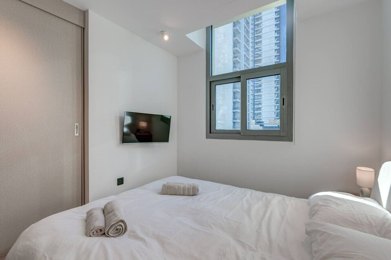 Une chambre d’hôtel avec un lit, un téléviseur fixé au mur, une fenêtre donnant sur des immeubles et une lampe de chevet.