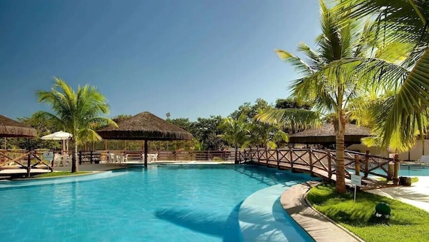 A swimming pool with a wooden deck, palm trees, and thatched-roof structures. - Apartamento Le Jardin  (Caldas Novas)