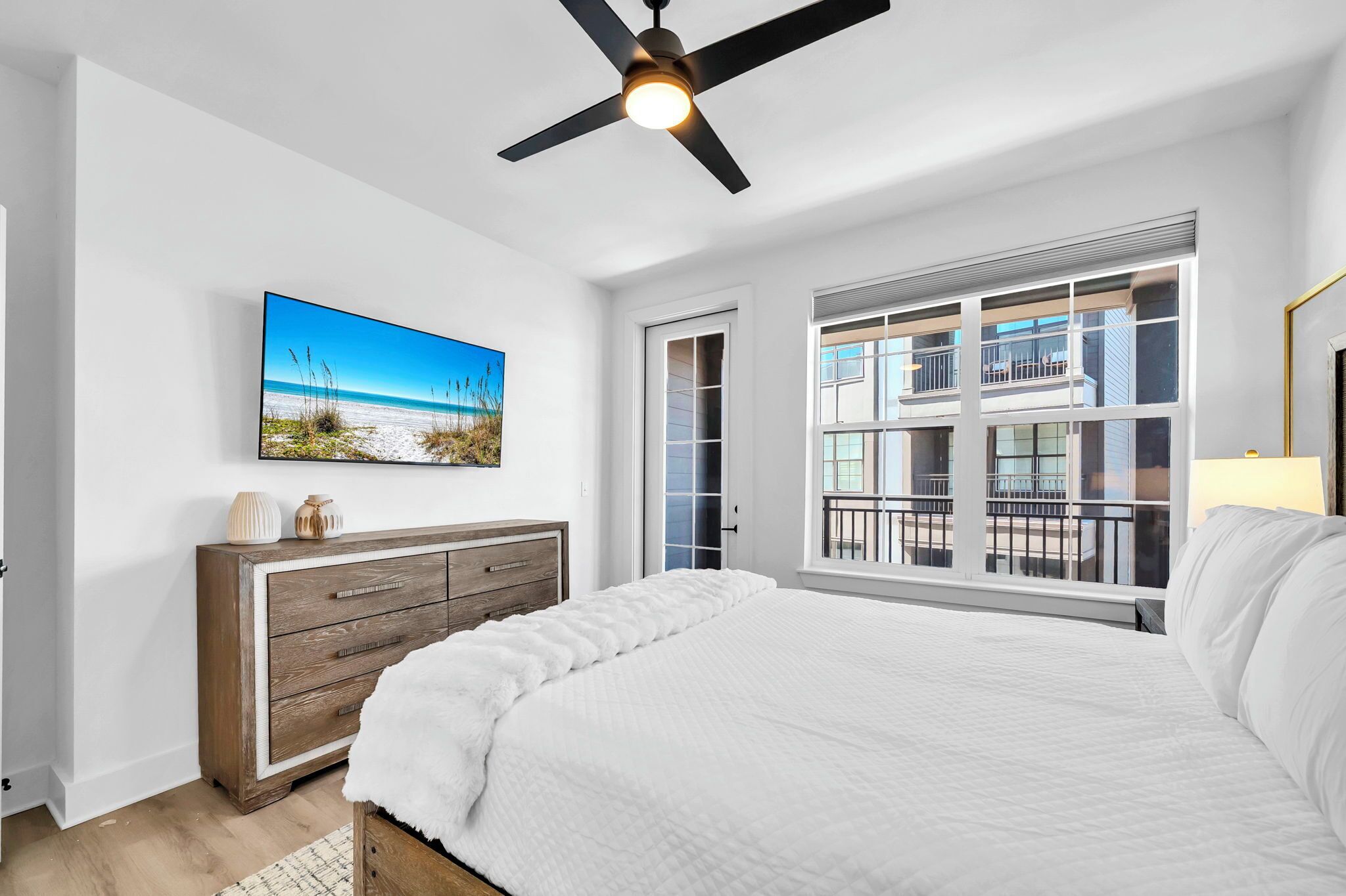 Une chambre à coucher avec un grand lit, un ventilateur de plafond, un tableau représentant une plage, une commode en bois et une fenêtre donnant sur un immeuble.