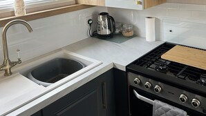 Een kleine keuken met witte kastjes, een RVS waterkoker, een gasfornuis en een raam met uitzicht op groen.