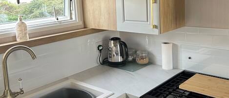 Une cuisine compacte avec des meubles blancs, une bouilloire en acier inoxydable, une cuisinière à gaz et une fenêtre donnant sur la verdure environnante.