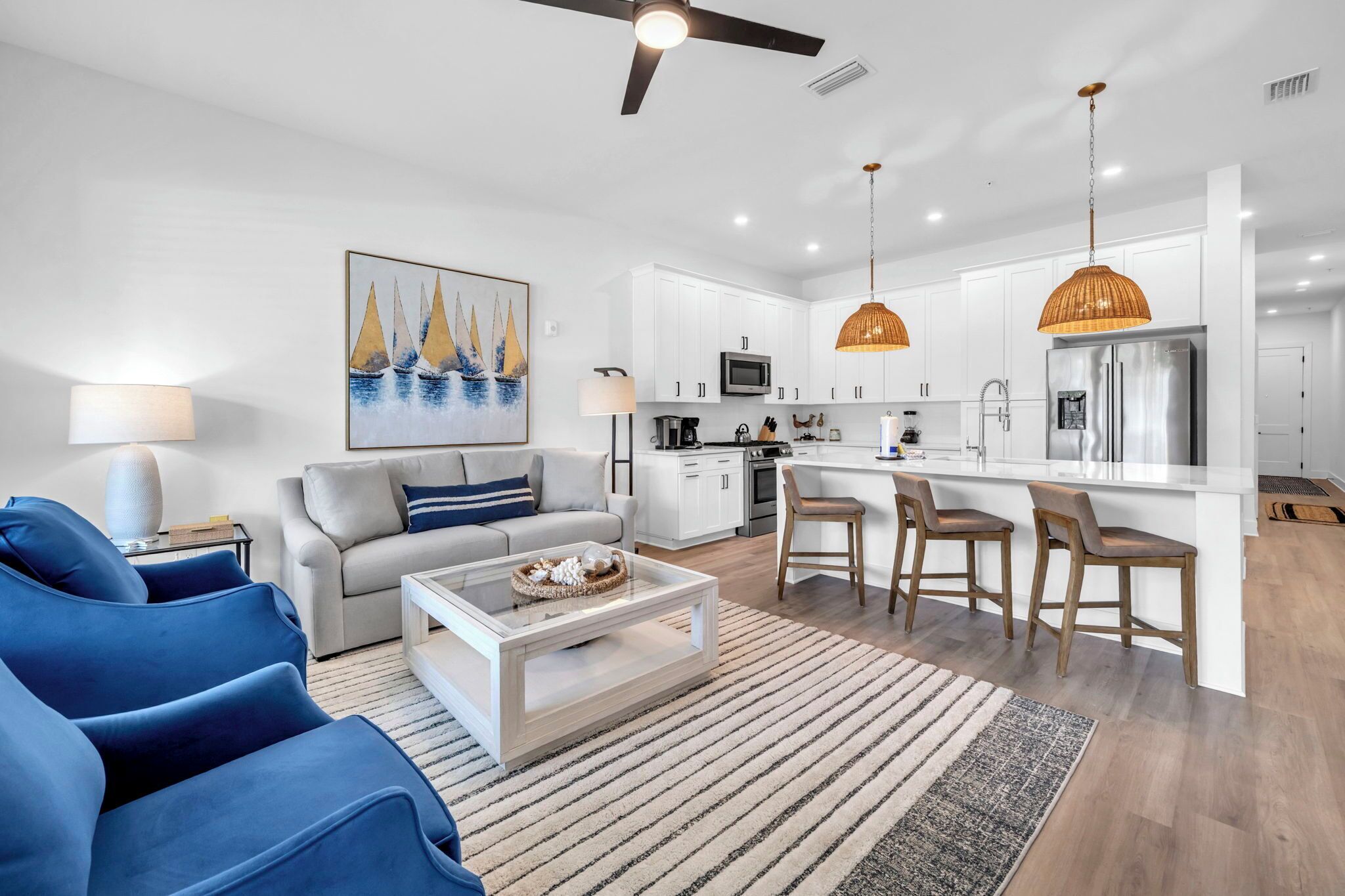 A modern living room with a sofa, coffee table, and blue chairs. A kitchen with white cabinets, a refrigerator, and pendant lights is visible.