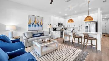 A modern living room with a sofa, coffee table, and blue chairs. A kitchen with white cabinets, a refrigerator, and pendant lights is visible.