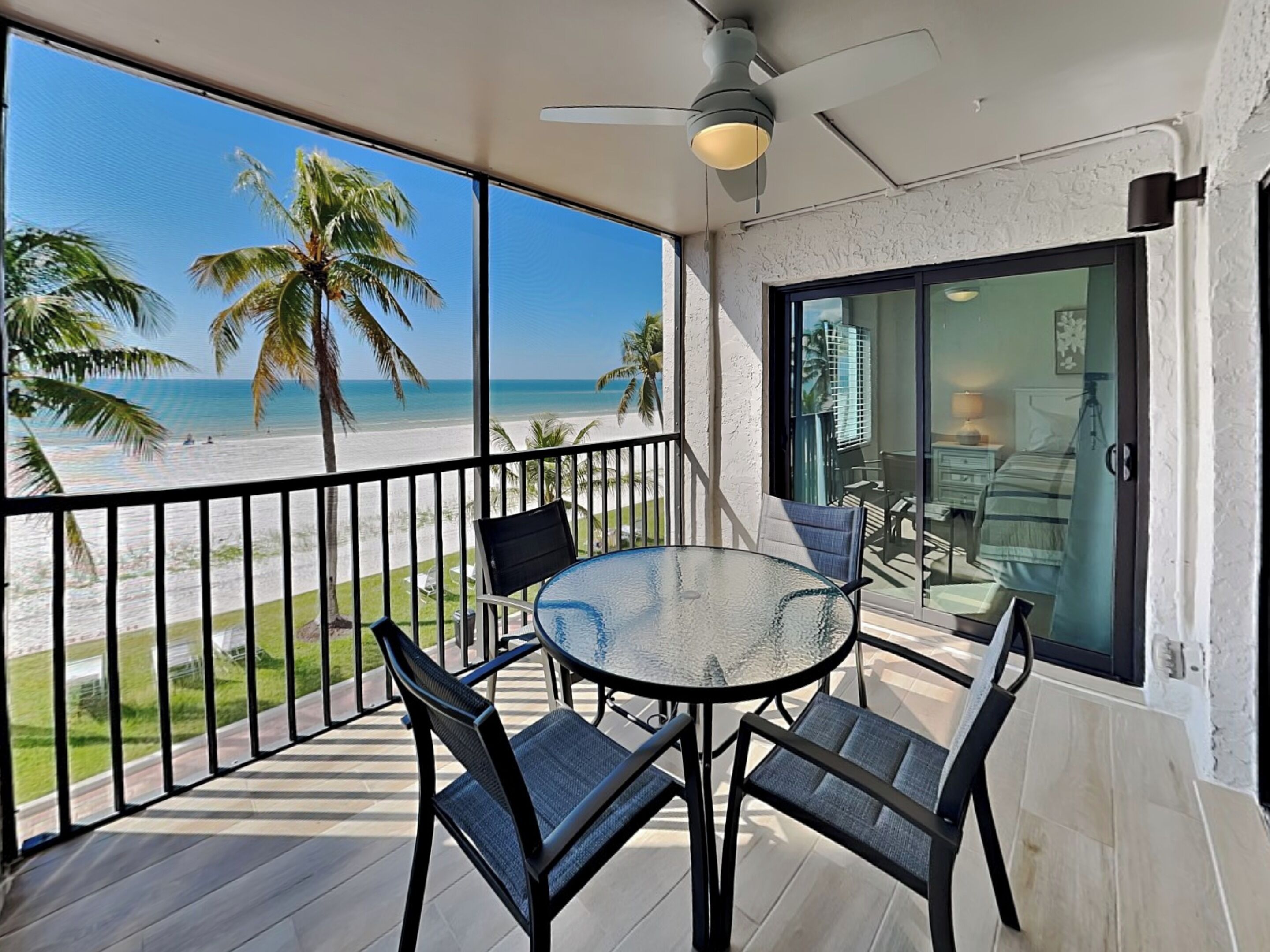 Ein Balkon mit Glastisch und Stühlen, einem Deckenventilator und Blick auf Strand und Palmen.