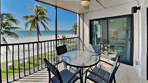 Uma varanda com mesa e cadeiras de vidro, um ventilador de teto e vista para a praia e palmeiras.