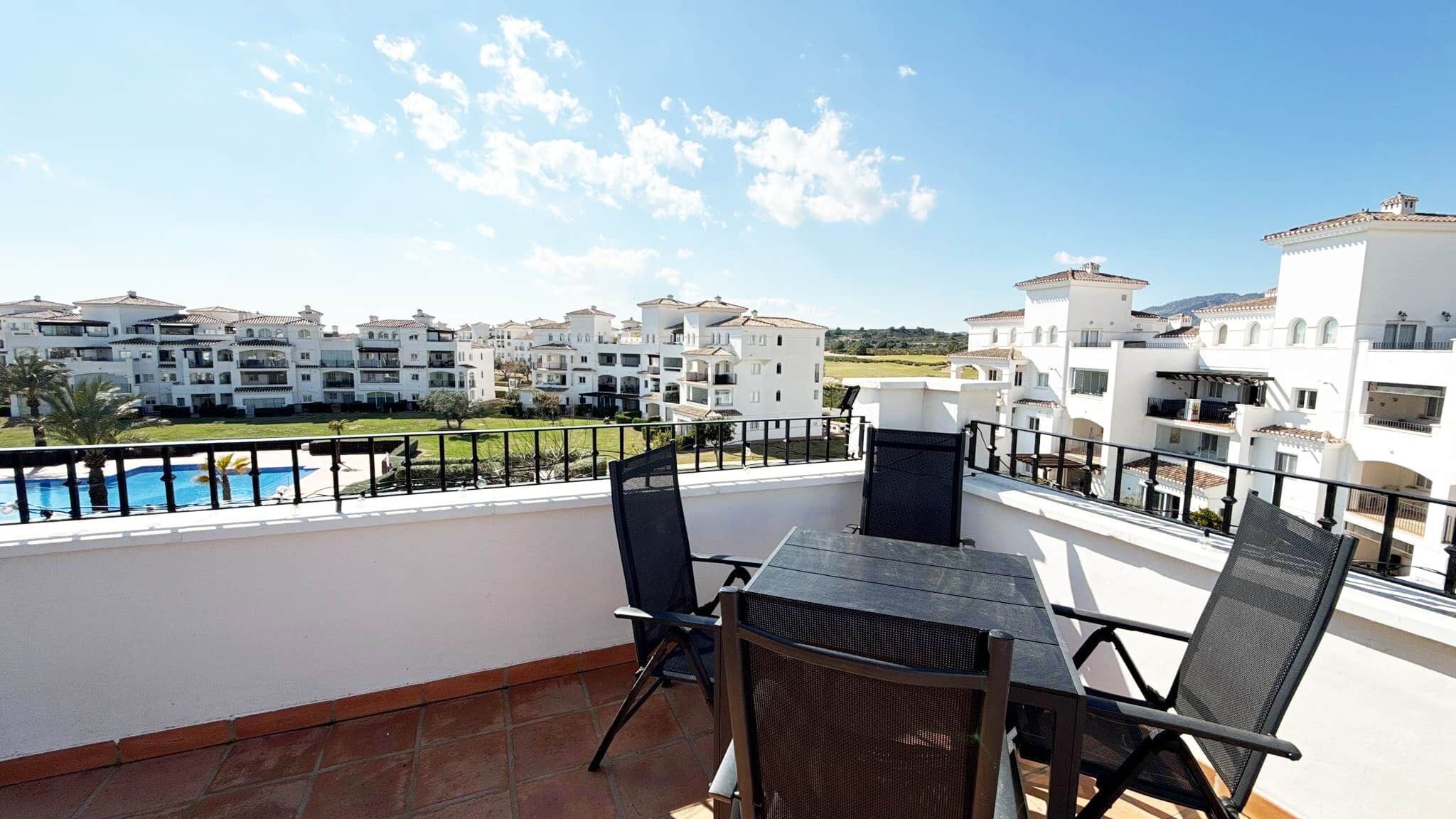A balcony with a table and chairs, overlooking a pool and white buildings.