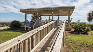 A wooden boardwalk leading to a covered area with a 'No Skateboard' sign, palm trees, and a view of the ocean. - Queen’s Court 2203 | Ocean Breeze Retreat (Emerald Isle)