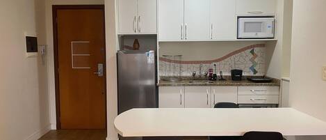 A compact kitchen with white cabinets, a stainless steel refrigerator, a microwave, and a dining area with a white table and two black chairs.