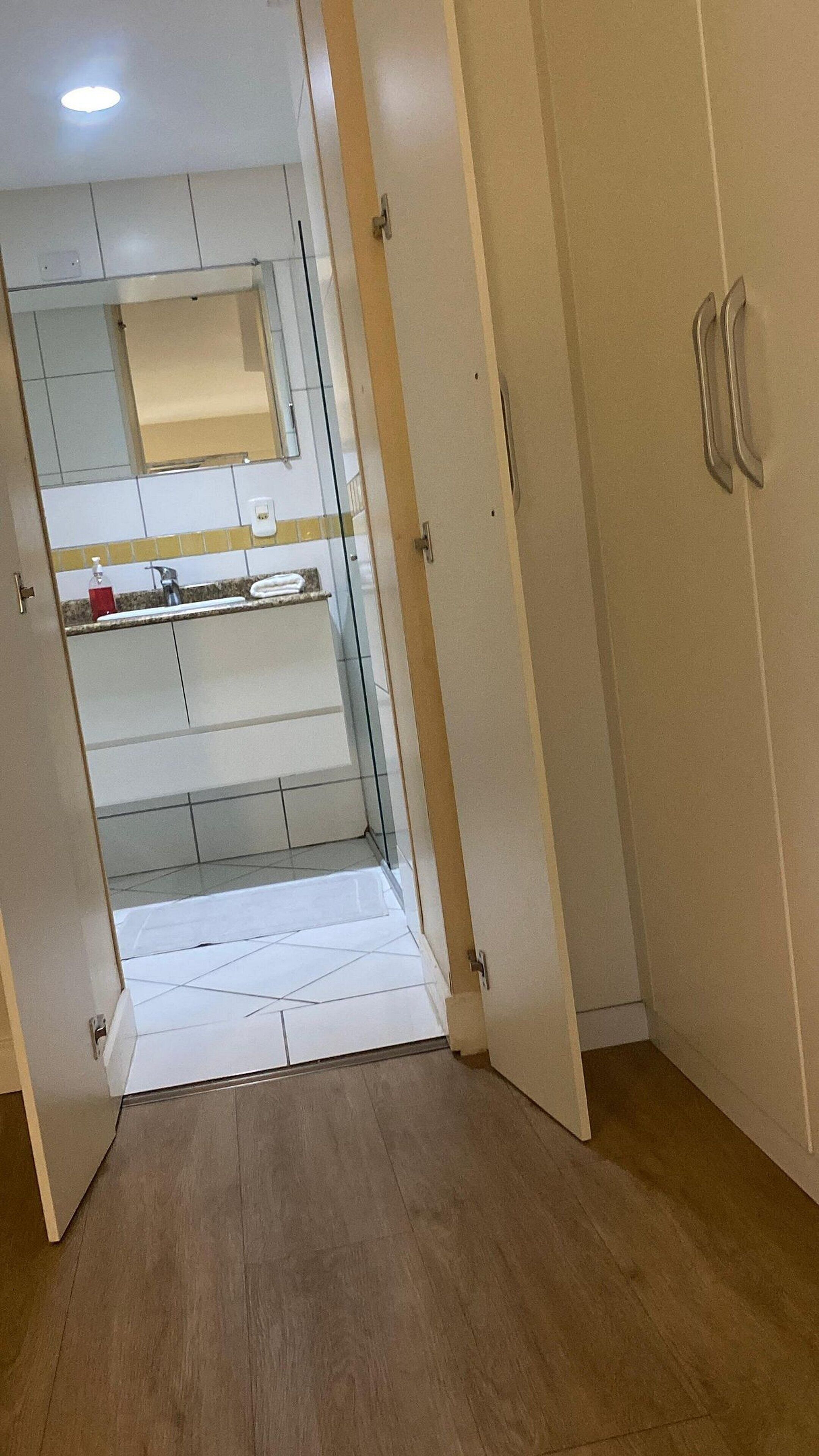 Una habitación con piso de madera, un armario blanco y una puerta de vidrio que da a un baño con espejo y toallero.