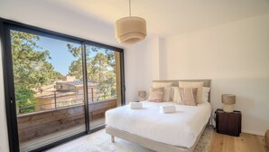 Een slaapkamer met een grote schuifpui, een bed met witte lakens, twee bedlampen en uitzicht op bomen en gebouwen.