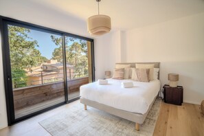 Une chambre avec une grande porte-fenêtre coulissante, un lit avec du linge de lit blanc, deux lampes de chevet et une vue sur les arbres et les bâtiments à l’extérieur.
