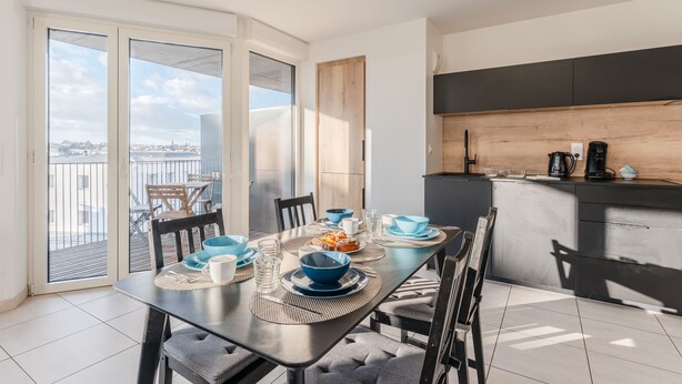 Zona pranzo con tavolo apparecchiato per quattro, cucina con piano di lavoro in legno e vista sulla città attraverso ampie finestre.