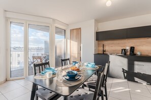 Un coin repas avec une table dressée pour quatre personnes, une cuisine dotée d’un plan de travail en bois et une vue sur la ville grâce à de grandes fenêtres.