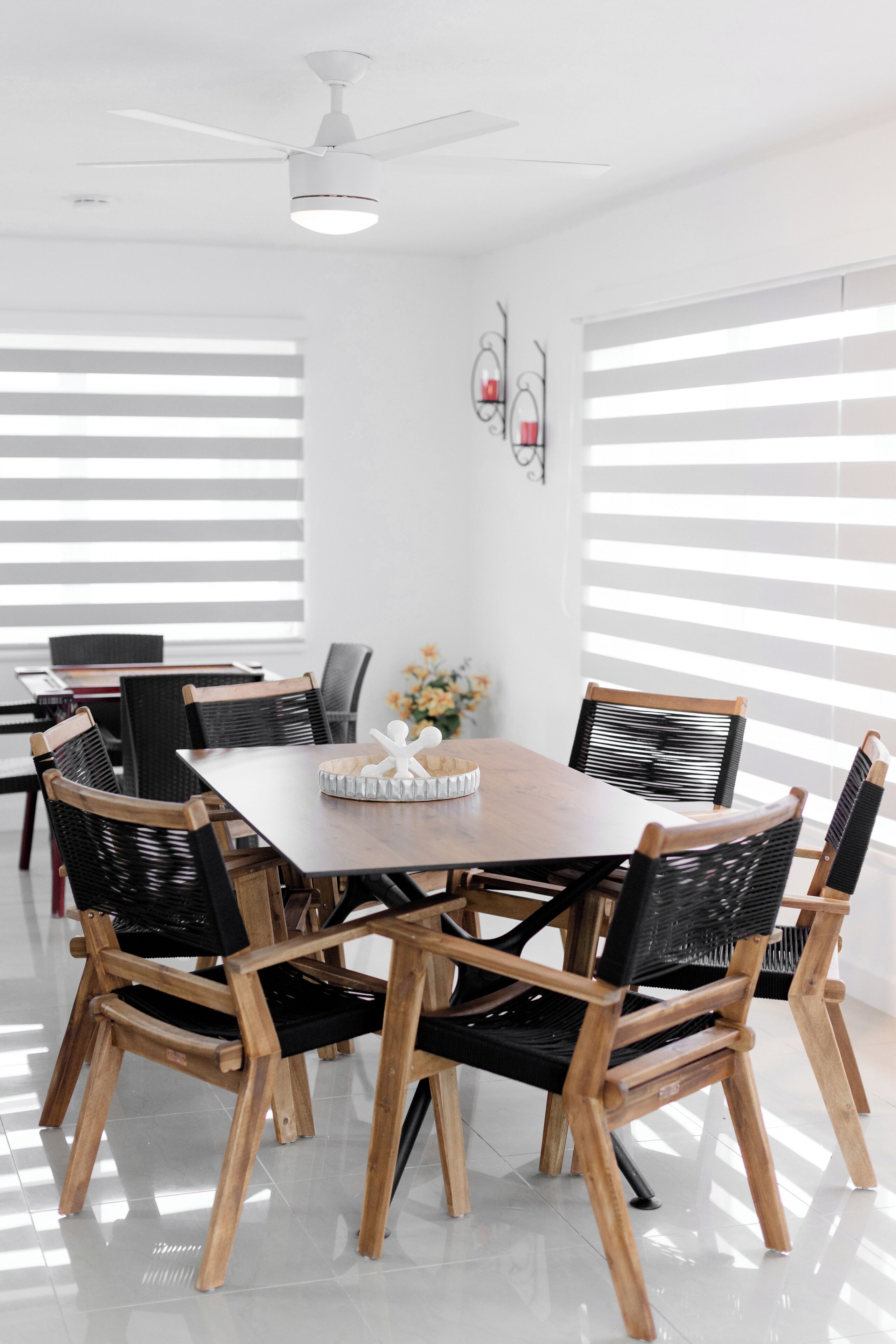 Ein Essbereich mit Holztisch und Stühlen, einem Deckenventilator und großen Fenstern mit Jalousien.