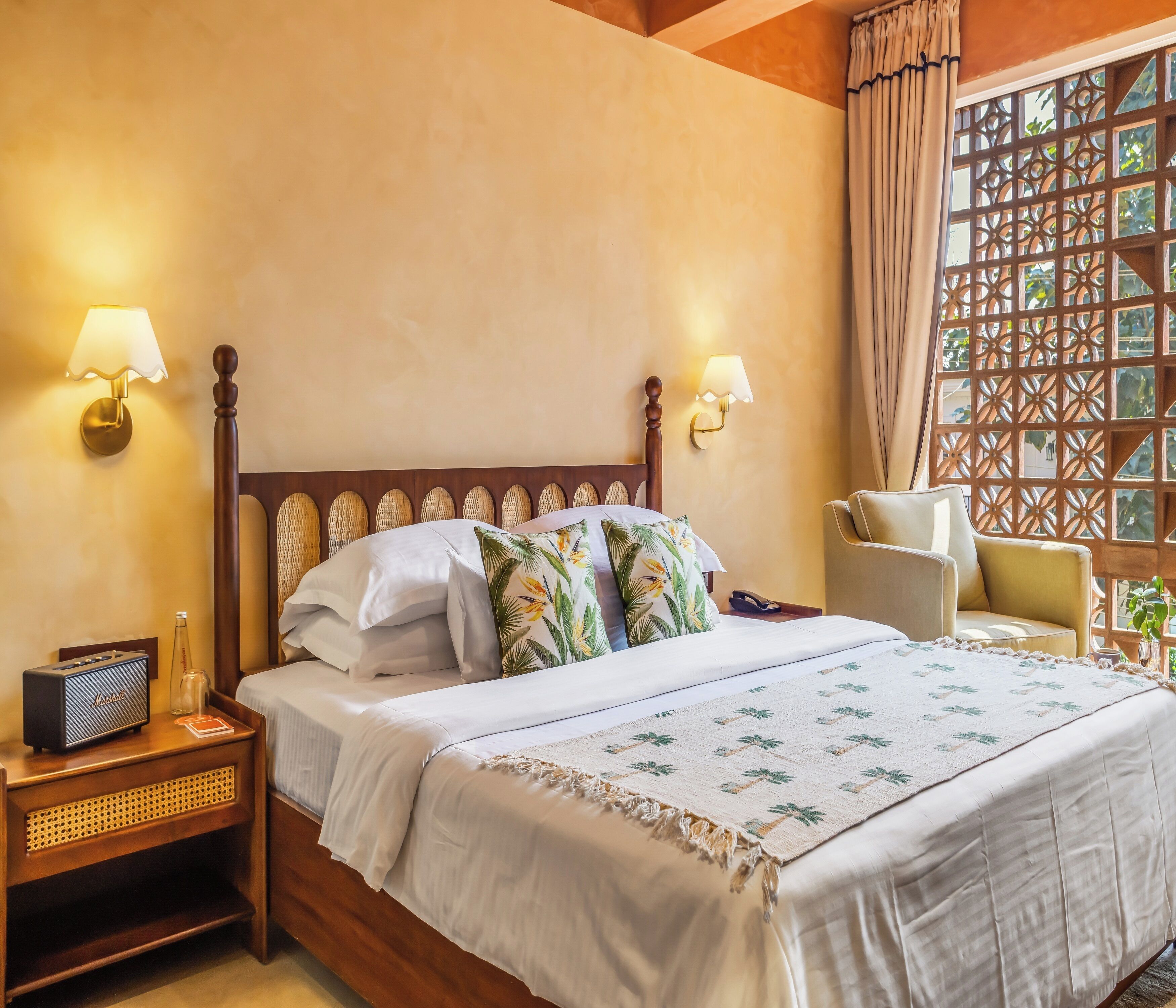 A neatly made bed with a white bedspread and two decorative pillows, a wooden nightstand with a radio, and a chair with a cushion.