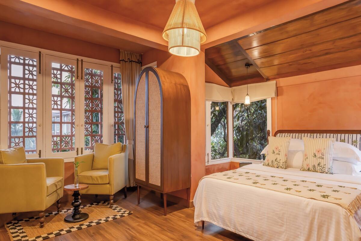 A bedroom with a wooden ceiling, a bed with floral bedding, a yellow armchair, and a large window with decorative shutters.