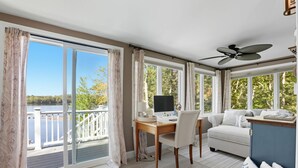 A room with a desk, chair, and a sofa. There is a large window with curtains, a ceiling fan, and a view of trees and a body of water. - Sawyer Lake NH, Lake & Ski – 4BR, all the amenities for Year-Round Adventure! (Gilmanton)