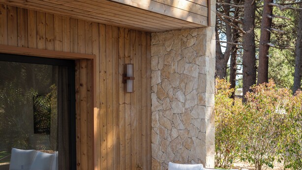 A wooden deck with wicker furniture, a stone wall, and a glass door leading to a covered area. - Villa Cala Rossa (Lecci)