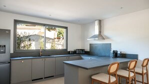 A modern kitchen with a large window, grey cabinets, a central island, and wooden bar stools. - Villa Cala Rossa (Lecci)