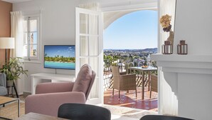 Una sala de estar con televisor, un sofá rosa y un balcón con mobiliario de mimbre.
