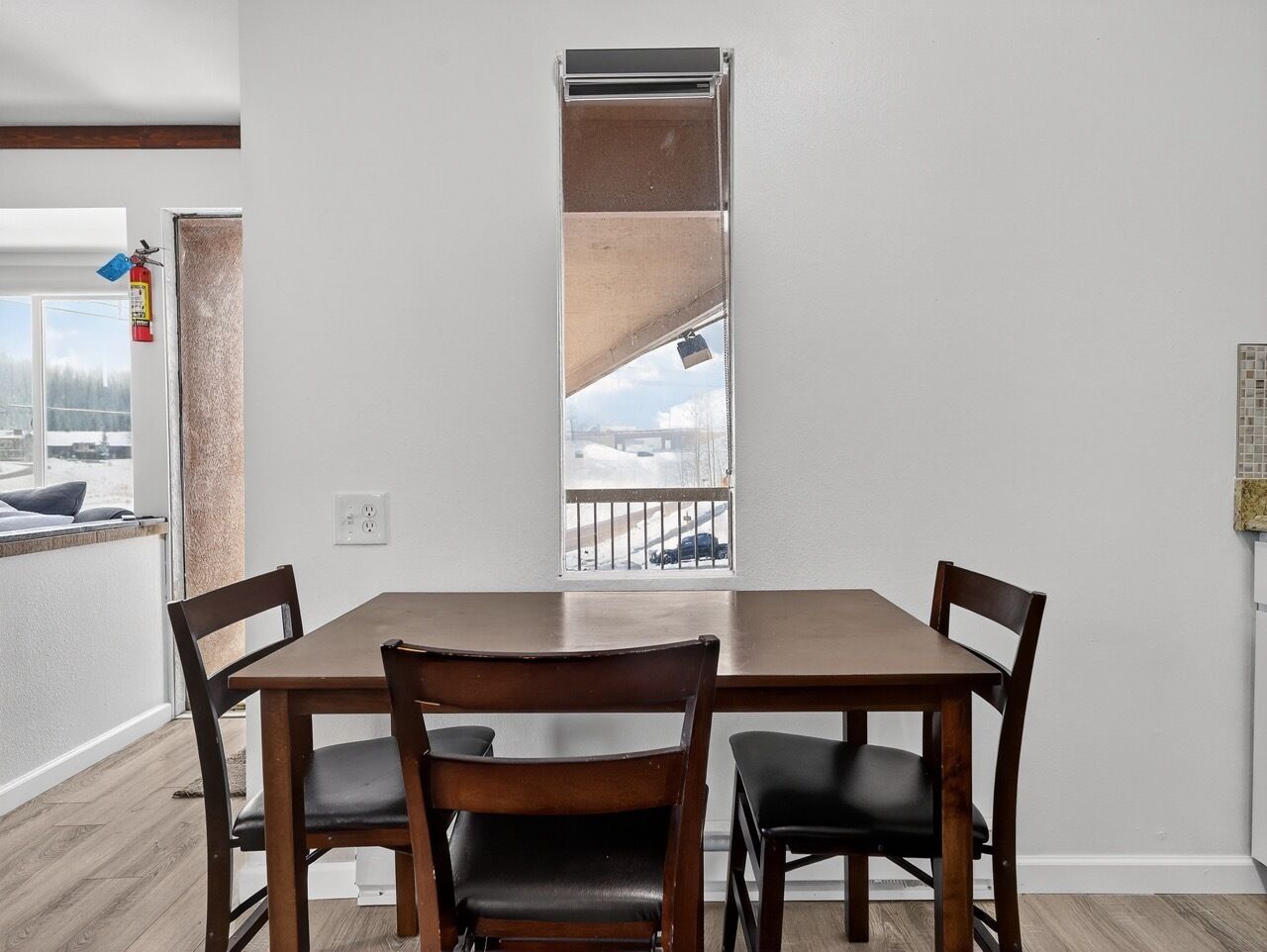 A dining area with a wooden table and four chairs, a window with a view of a snowy landscape, and a fire extinguisher mounted on the wall.