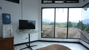A room with a large window, a television on a stand, a water dispenser, and a woven floor mat. - Villa Agape Near Puncak By ARBA (Cipanas)