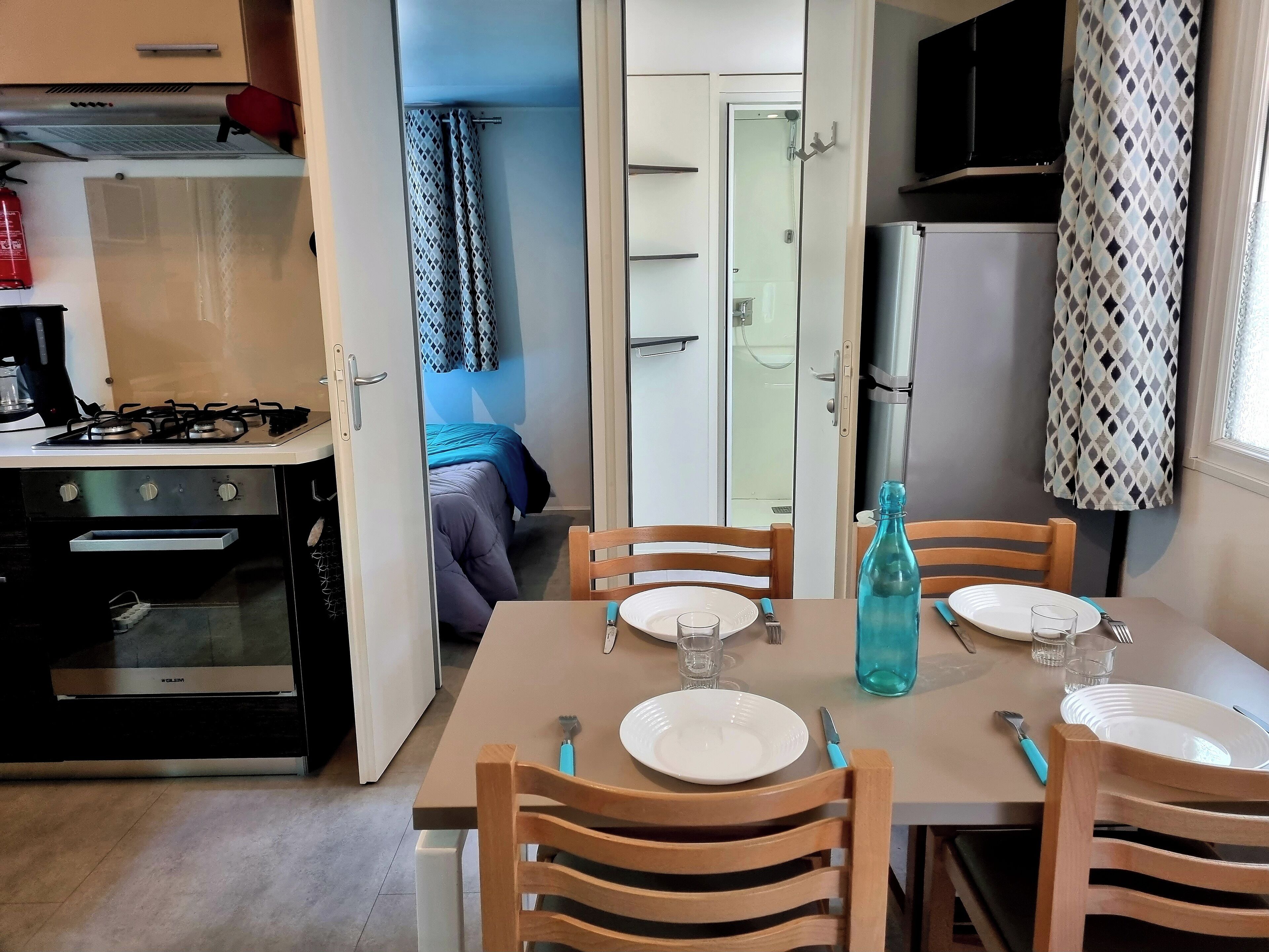 A compact kitchen with a gas stove, oven, and a microwave. A dining table with chairs is set up in the center, with plates, cutlery, and a blue bottle on it.