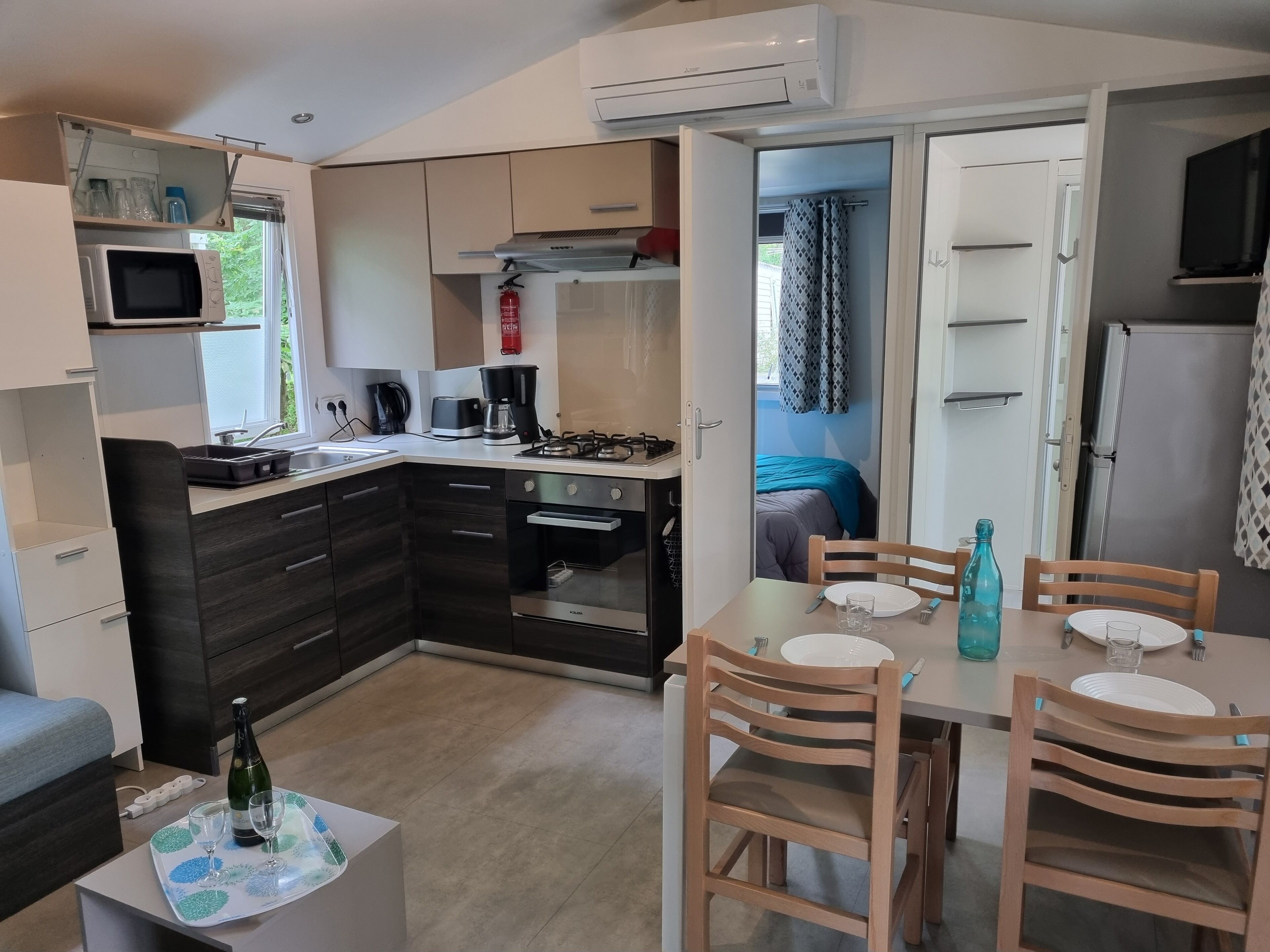 A compact kitchen with modern appliances, a dining area with wooden chairs, and a visible bed in the adjacent room.